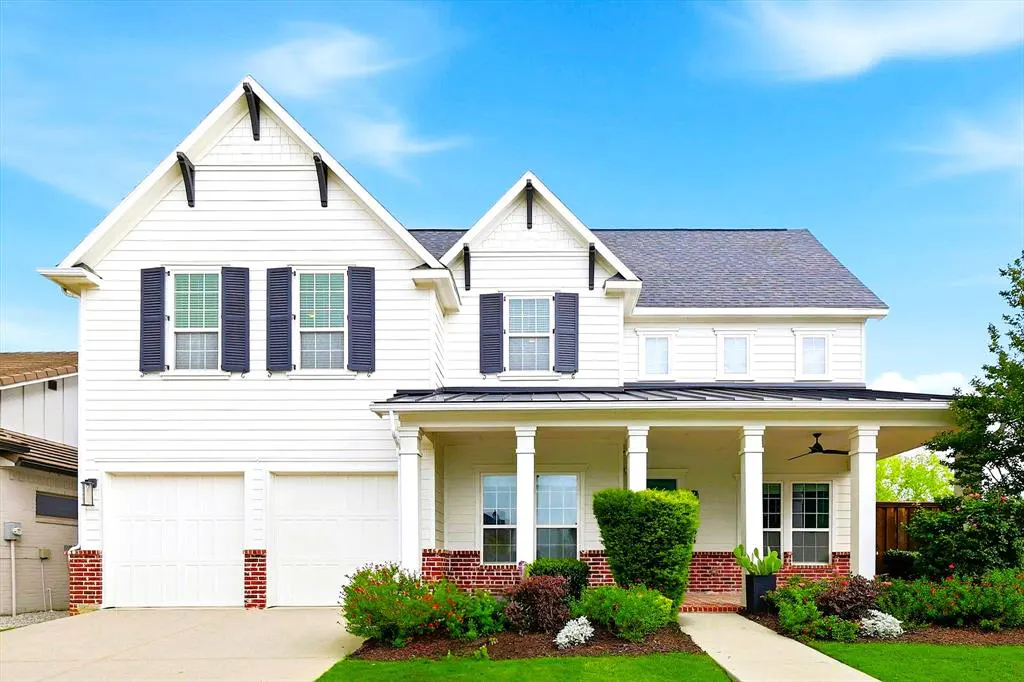 Two-story white house with black shutters, gray roof, and a covered porch with white columns. Landscaped yard with green grass and colorful bushes.