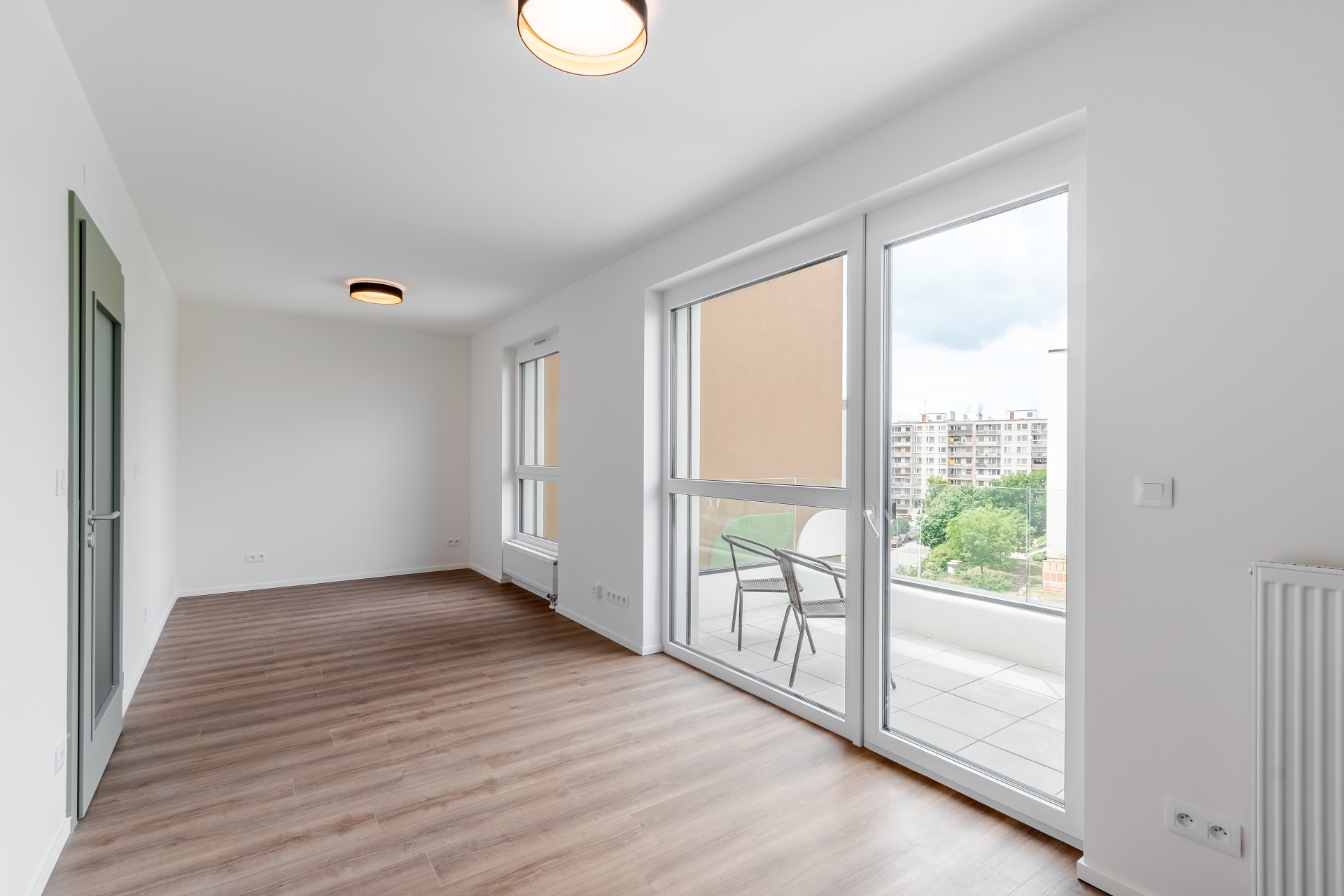 Bright, empty room with wood-look floors, white walls, and a balcony with two chairs overlooking a city view.