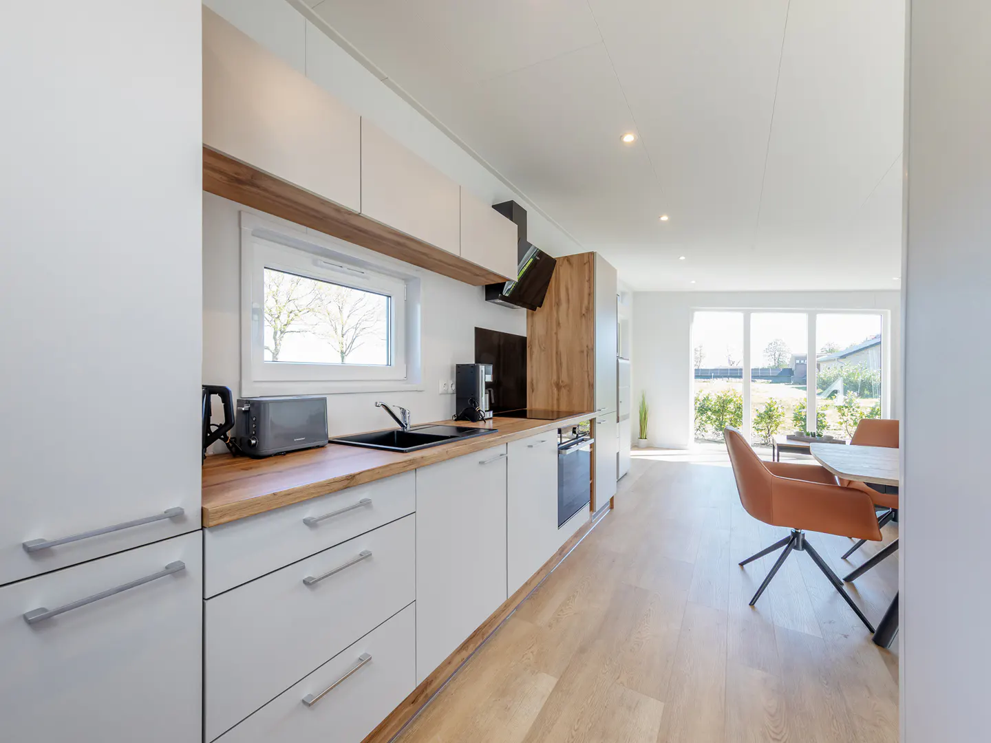 Bright, modern kitchen with white cabinets, wood countertop, and stainless steel appliances. Dining area with brown chairs and large windows in the background.