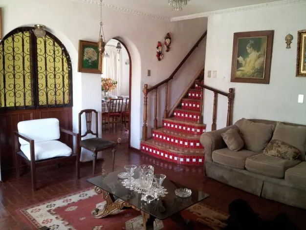 Living room with a brown sofa, glass table, and red-and-white tiled stairs. Arched doorways lead to other rooms.