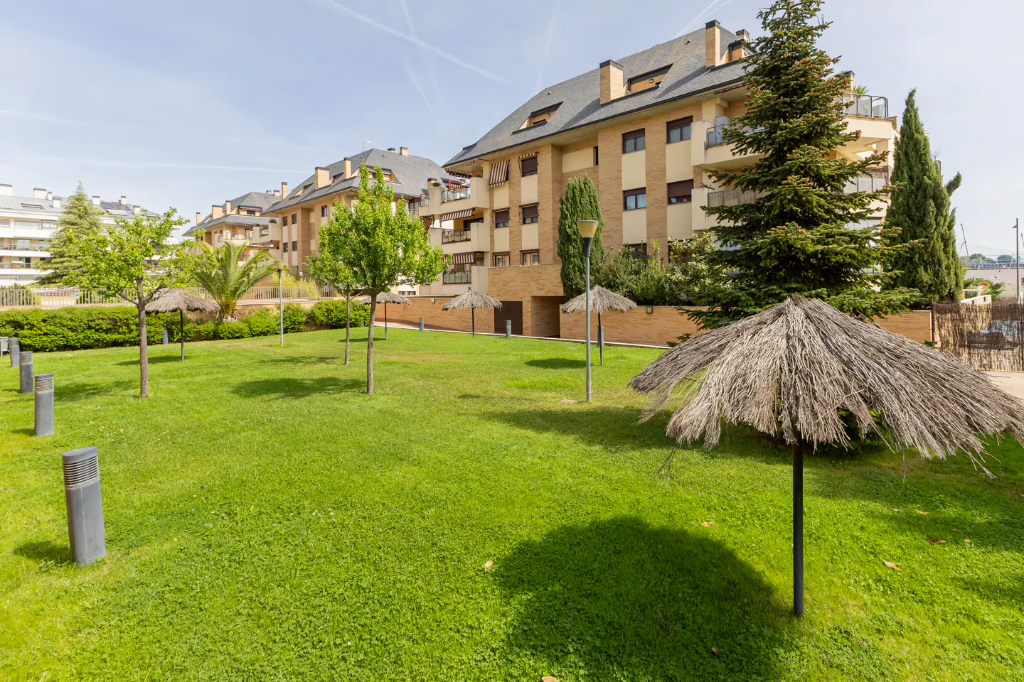 Apartment complex with a green lawn, trees, and straw umbrellas on a sunny day.