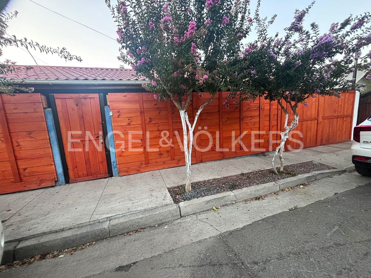 A long, orange wooden fence with two trees in front, one with pink flowers. The Engel & Völkers logo is on the fence.