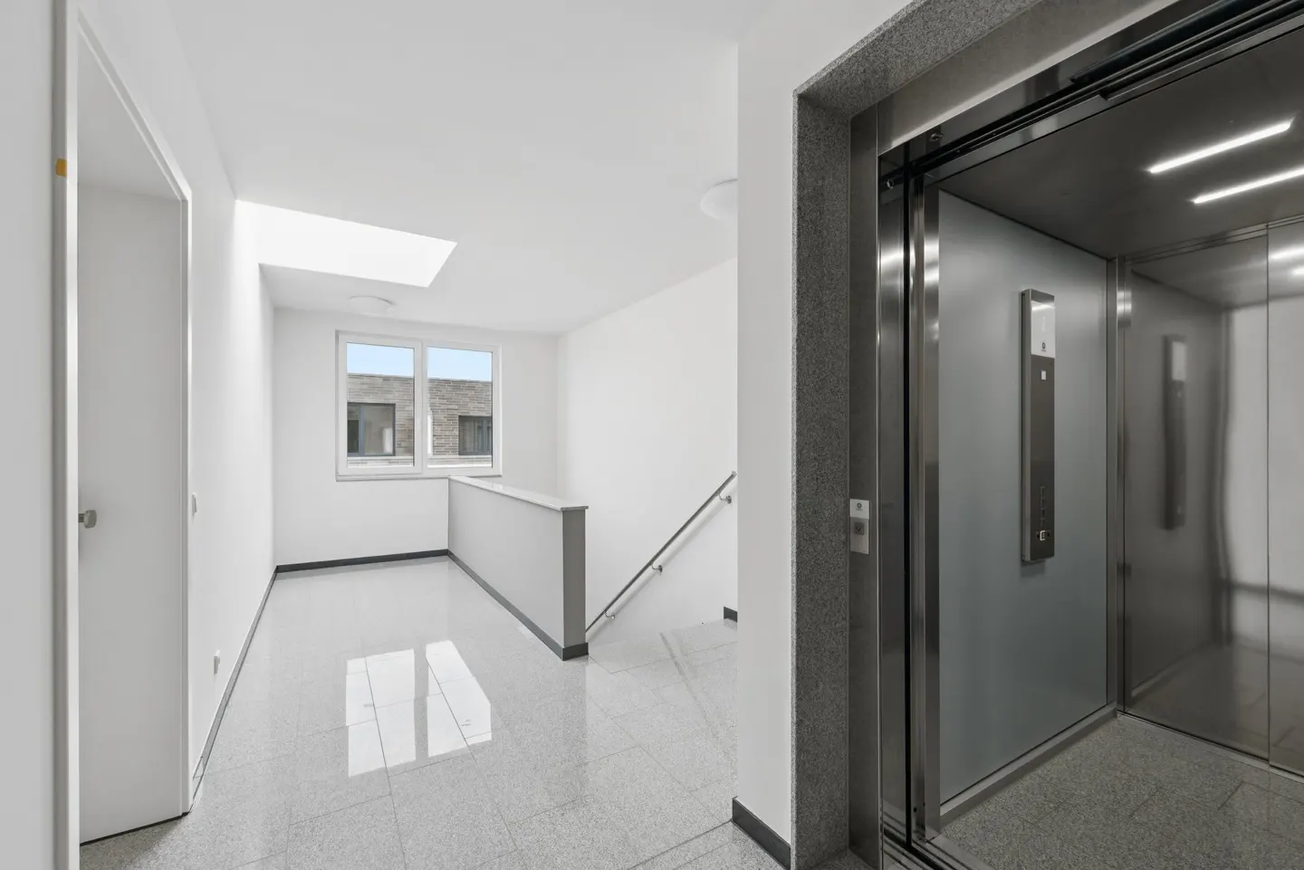 Bright, modern hallway with gray tile floor, white walls, stairs, and a stainless steel elevator with open doors.