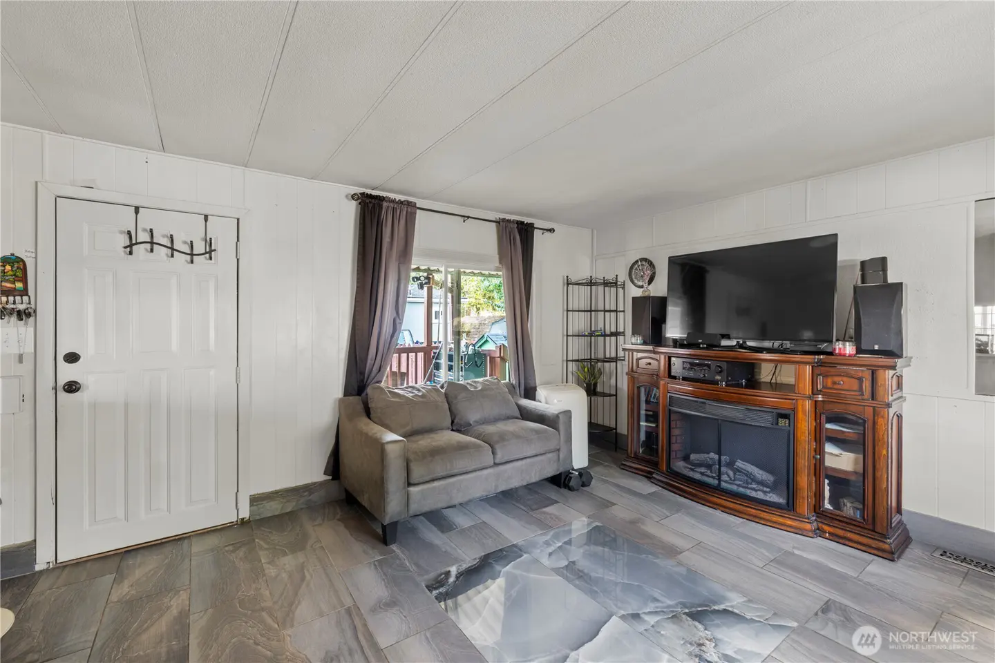 Living room with gray sofa, wood-look tile floor, and white walls. A TV sits atop a wooden fireplace console.