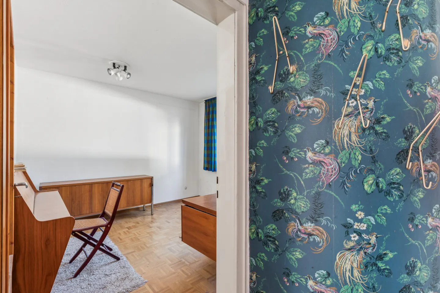 View through a doorway into a room with wood furniture and parquet floors, beside a wall with blue bird-pattern wallpaper.