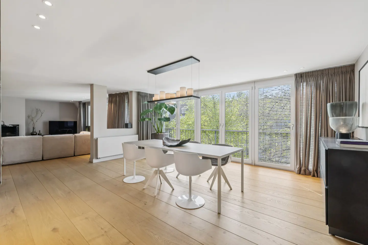 Bright dining area with wood floors, white table and chairs, and large windows with blinds and curtains. A black cabinet is on the right.