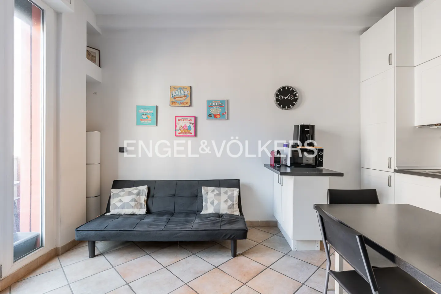 A compact apartment interior with a black futon, kitchen appliances, and a dining table. The walls are white, decorated with colorful food-themed art.