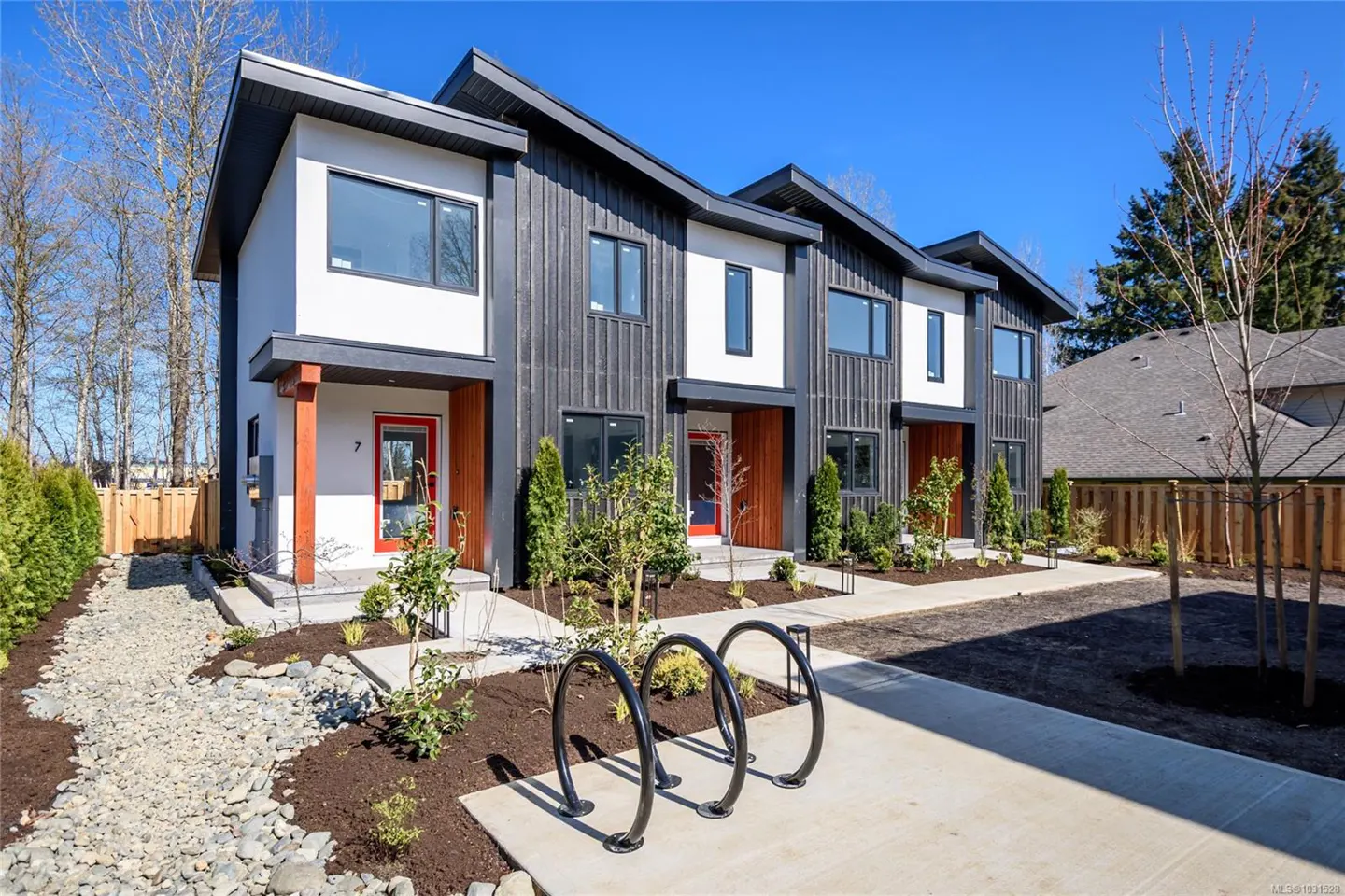Modern townhouses with white and dark siding, red doors, and black trim under a blue sky. A bike rack sits in front.