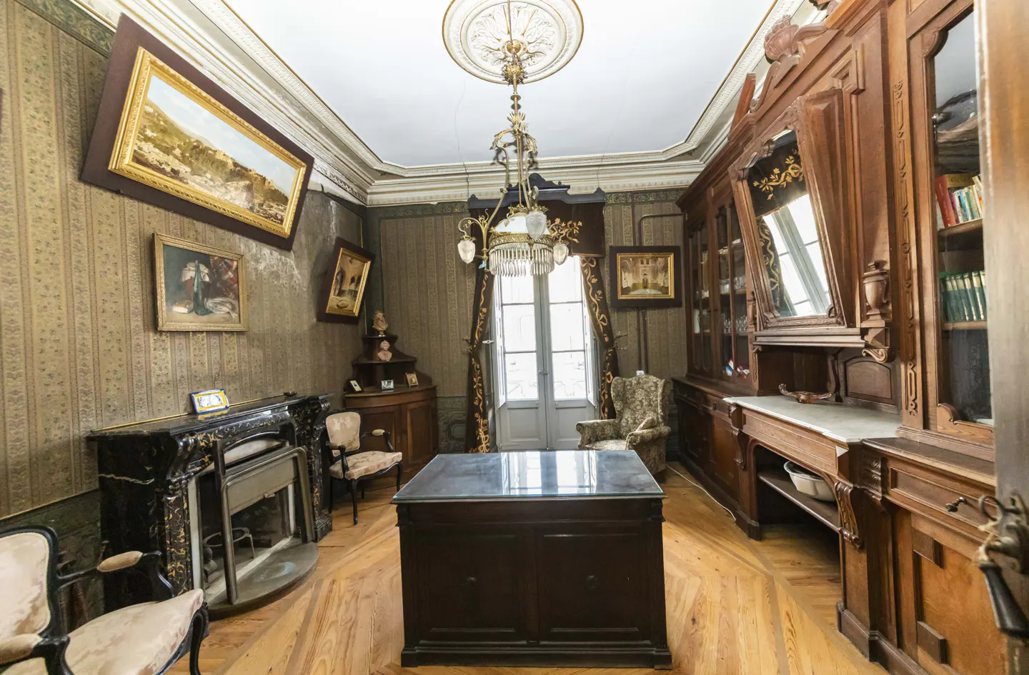 Ornate room with wood floors, patterned wallpaper, and a large wooden desk. A fireplace, paintings, and a chandelier add to the room's character.