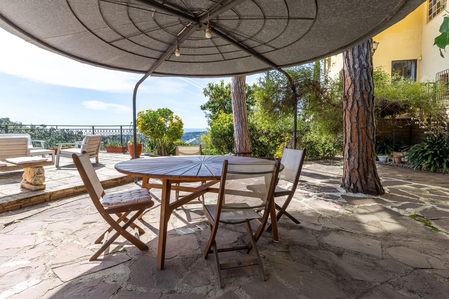 Outdoor patio with a round wooden table and chairs under a metal canopy. Stone flooring, trees, and a view of the landscape in the background.