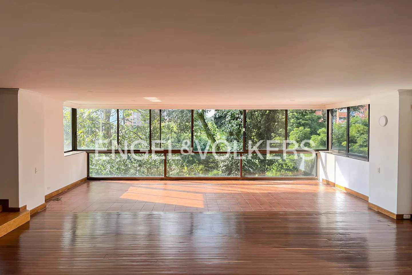 Empty living room with hardwood floors and a wall of windows overlooking lush greenery. Engel & Volkers logo visible on the glass.