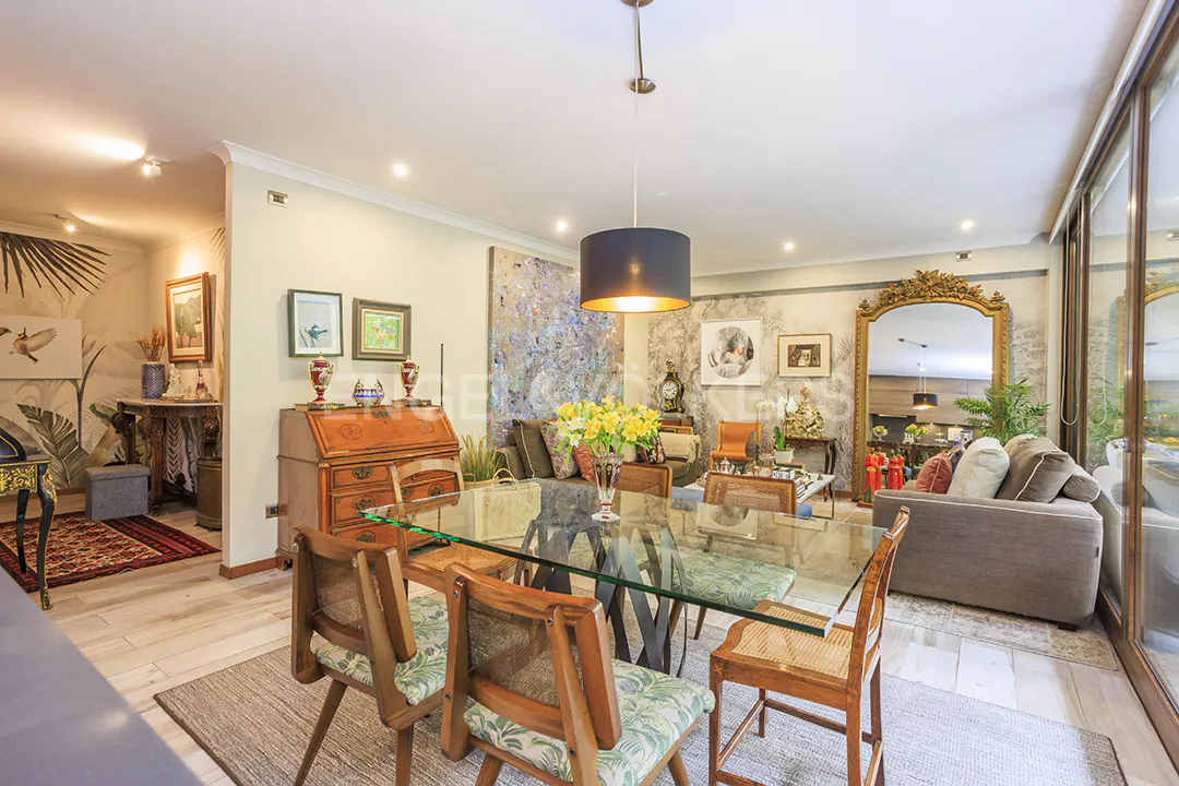 A bright living room with a glass table, wooden chairs, and a brown sofa. A large mirror and artwork decorate the walls.