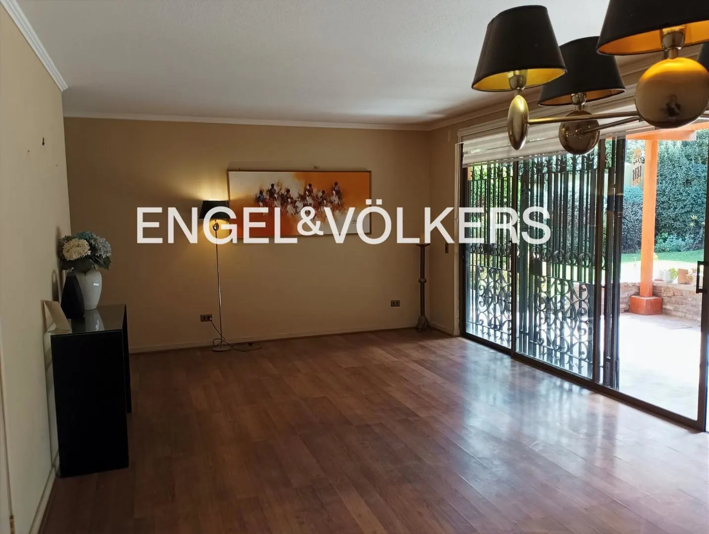 A living room with hardwood floors, tan walls, and a sliding glass door to a patio. A painting hangs above a floor lamp.