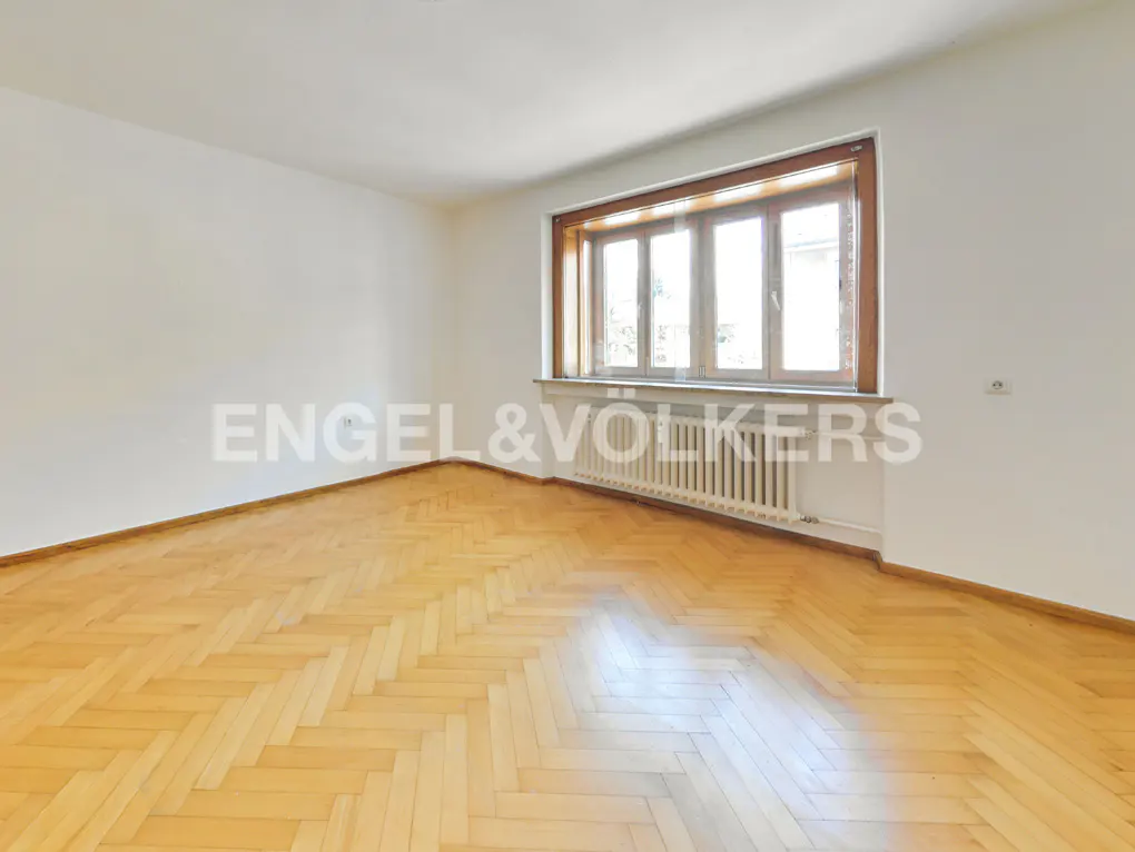Empty room with white walls, light wood herringbone floors, and a large window with a wooden frame and white radiator.