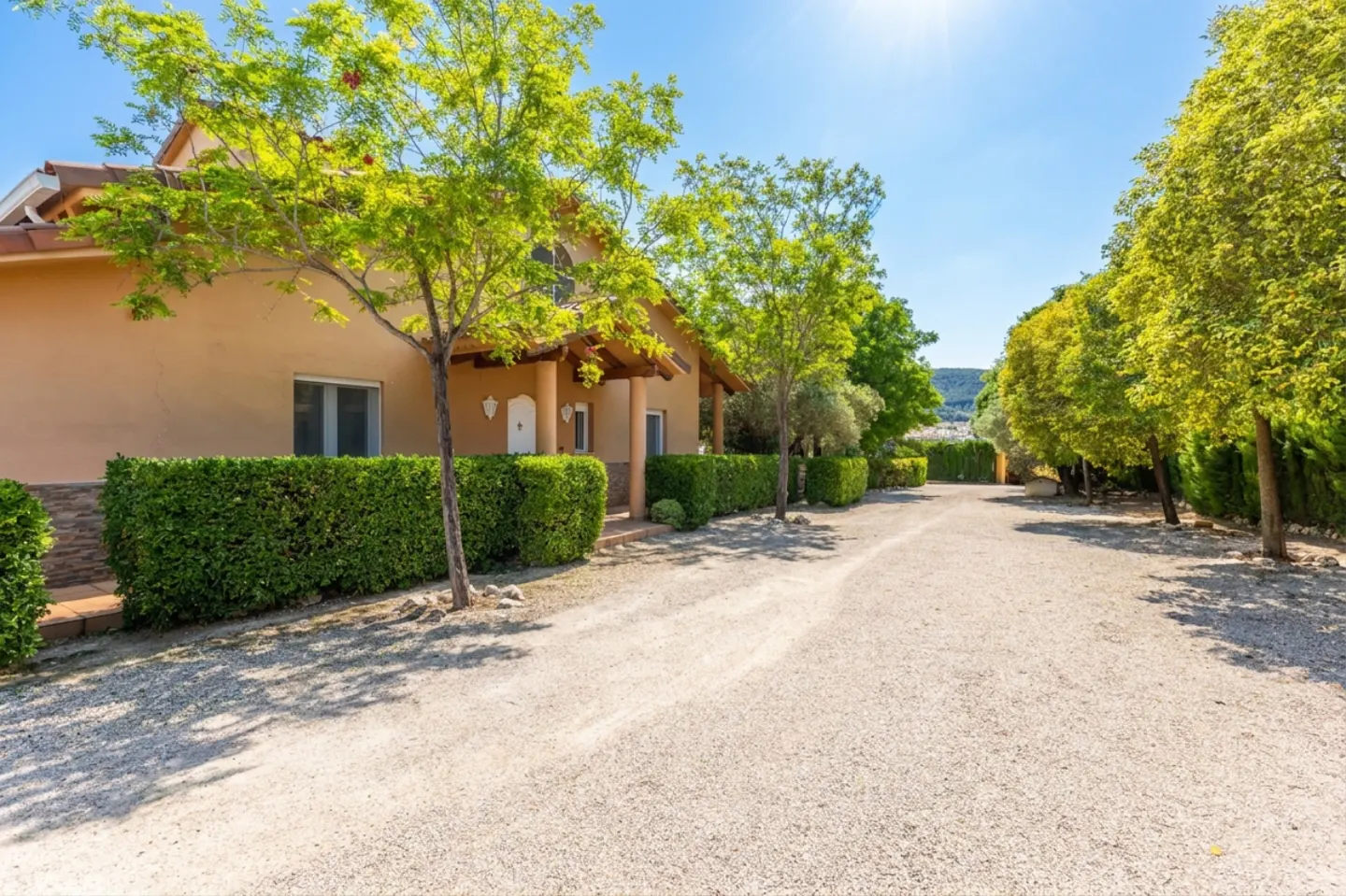 Exterior of a tan house with a gravel driveway, green trees, and trimmed hedges under a blue sky.