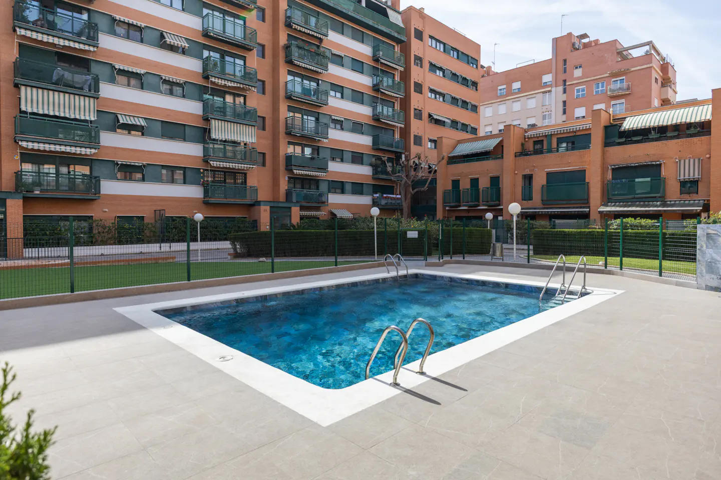 Outdoor pool with blue water and metal ladders, surrounded by gray tile. Orange brick apartment buildings in the background.