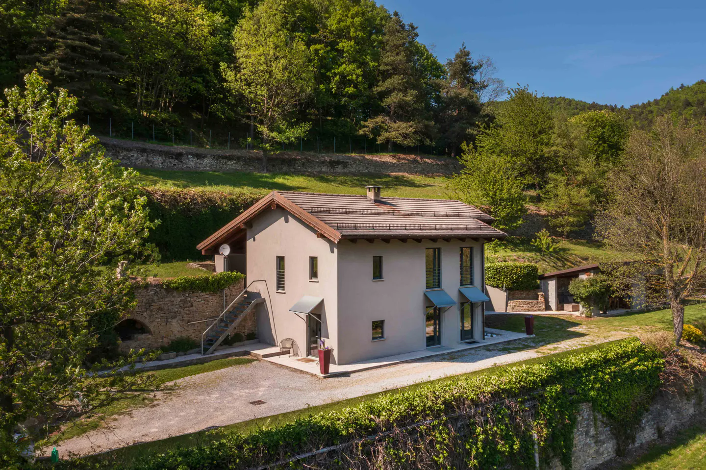 Two-story gray house with a brown roof, surrounded by green trees and a stone wall covered in ivy.