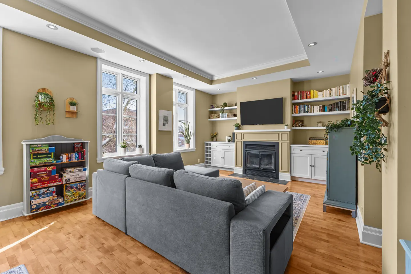 Living room with hardwood floors, a gray sectional sofa, and a fireplace with a TV above it. There are bookshelves and cabinets on either side of the fireplace. A bookcase filled with board games is on the left.