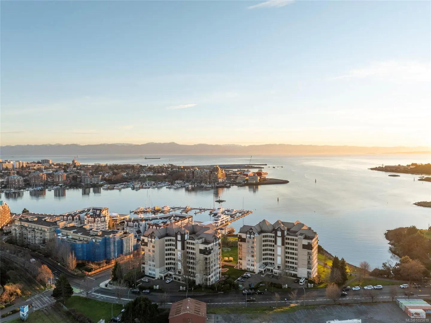 Aerial view of Victoria, BC, with harbor, buildings, and mountains in the distance under a blue sky with golden sunlight.