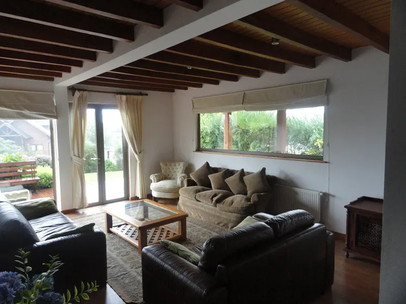 Living room with brown leather sofas, a tan sofa, and a glass coffee table on a patterned rug. A garden is visible through the windows.
