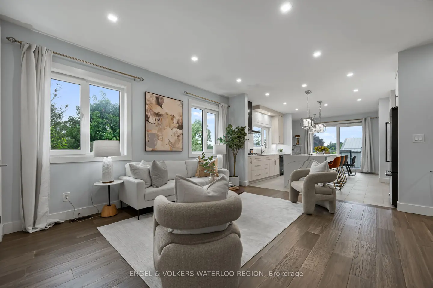 Open-concept living room and kitchen with hardwood floors, white sofa, and beige armchairs. Large windows offer natural light.