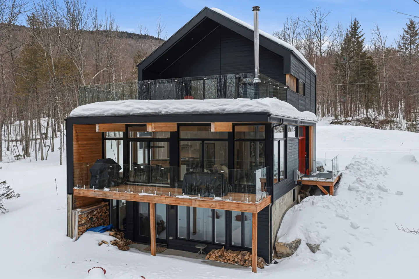 A modern black house with a glass balcony is surrounded by snow and trees.