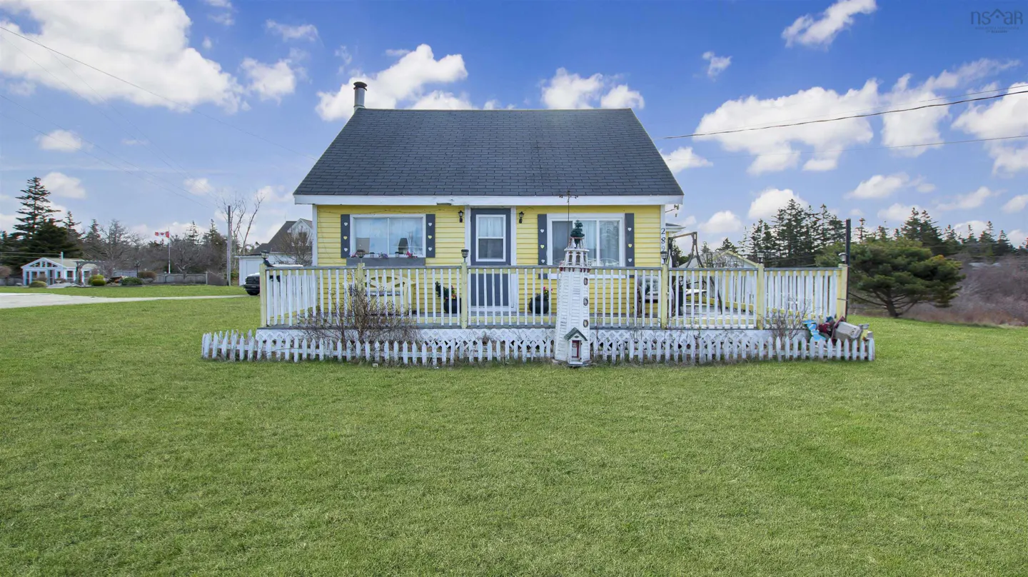 A yellow cottage with a gray roof, white trim, and a white picket fence sits on a green lawn under a blue sky.