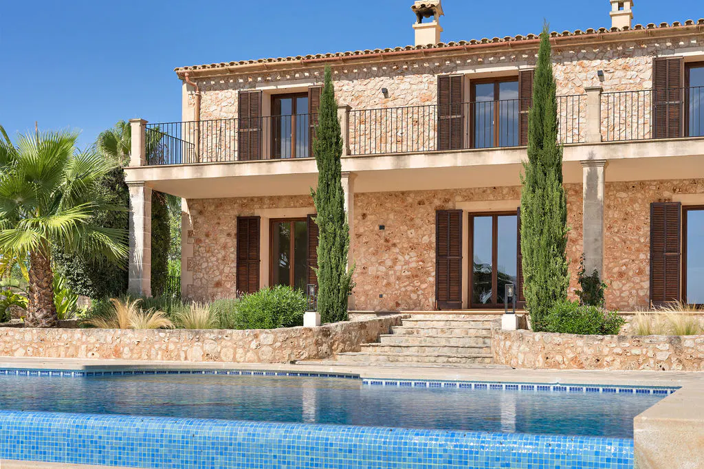 Two-story stone house with a blue tiled pool in the foreground on a sunny day. Balcony with dark railings and brown shutters.
