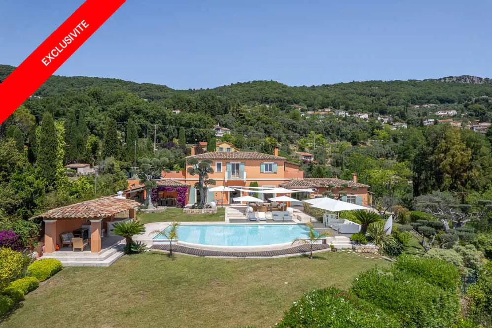 Orange villa with a pool, patio, and covered terrace, set in a lush green landscape under a clear blue sky.