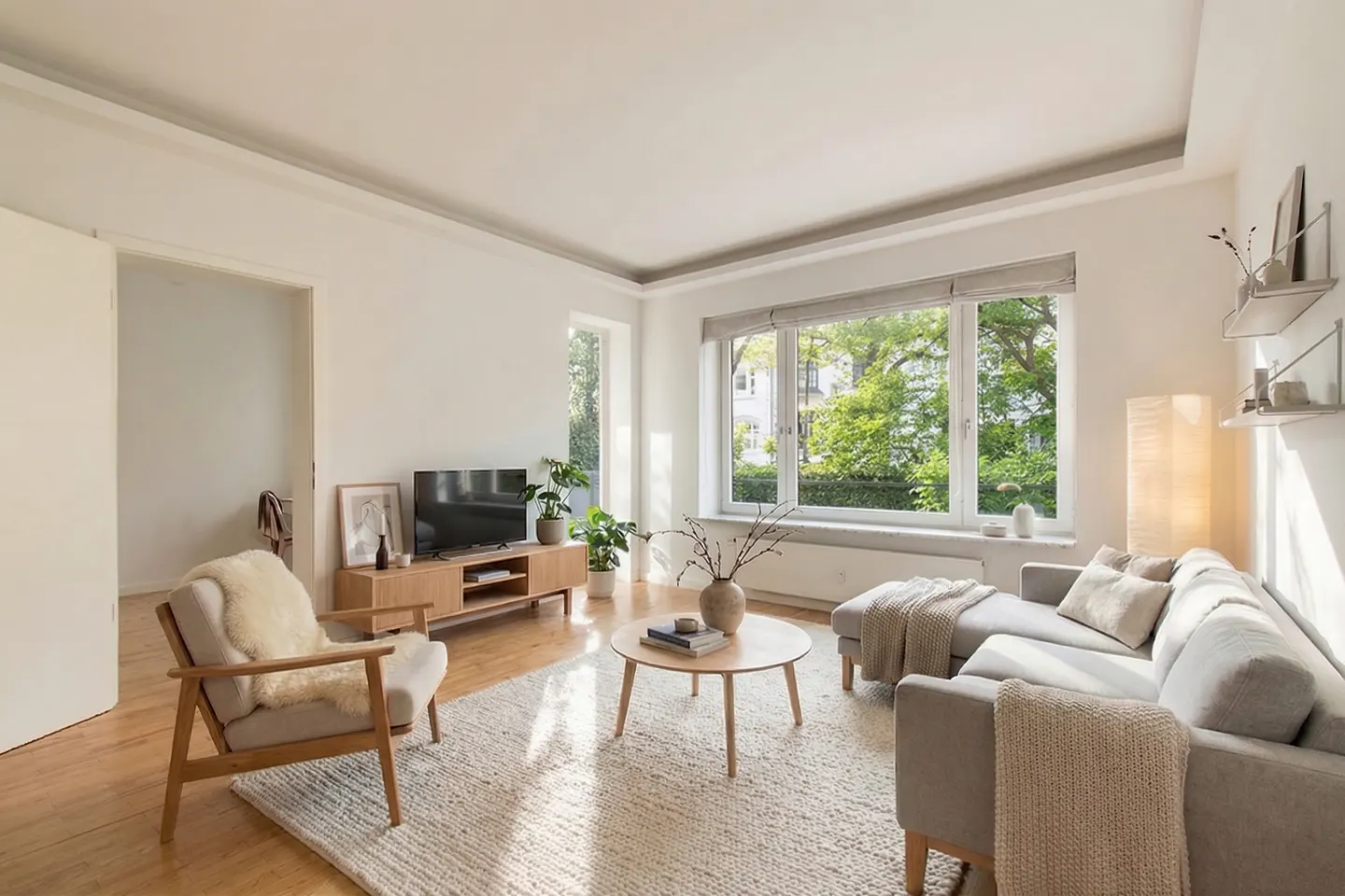 Bright living room with a gray sectional sofa, wood floors, and a large window overlooking green trees. A round coffee table sits on a textured rug.