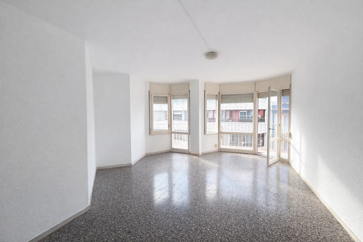 Empty, bright room with white walls, speckled grey flooring, and large windows leading to a balcony.