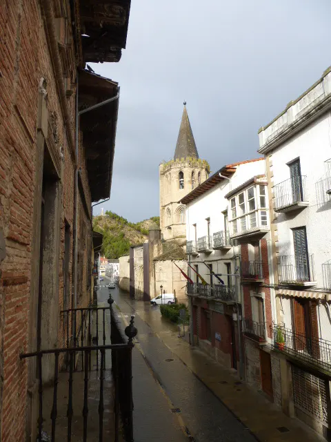 View of a wet street in a European town, lined with buildings and a church steeple in the background.