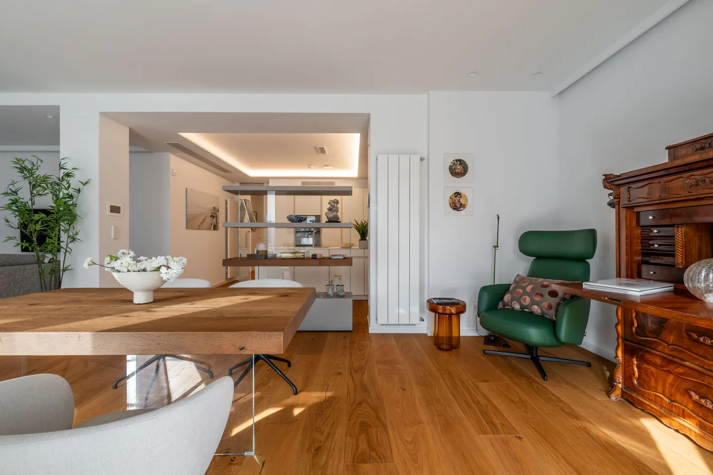 Bright, modern living room with wood floors, a dining table with white chairs, and a green leather armchair next to a dark wood antique cabinet.