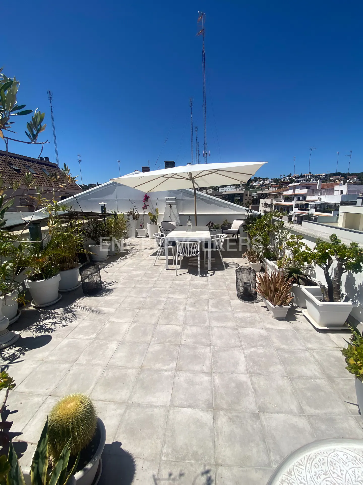 Rooftop patio with white table, chairs, and sun umbrella. Potted plants line the perimeter, with city buildings visible in the background under a blue sky.