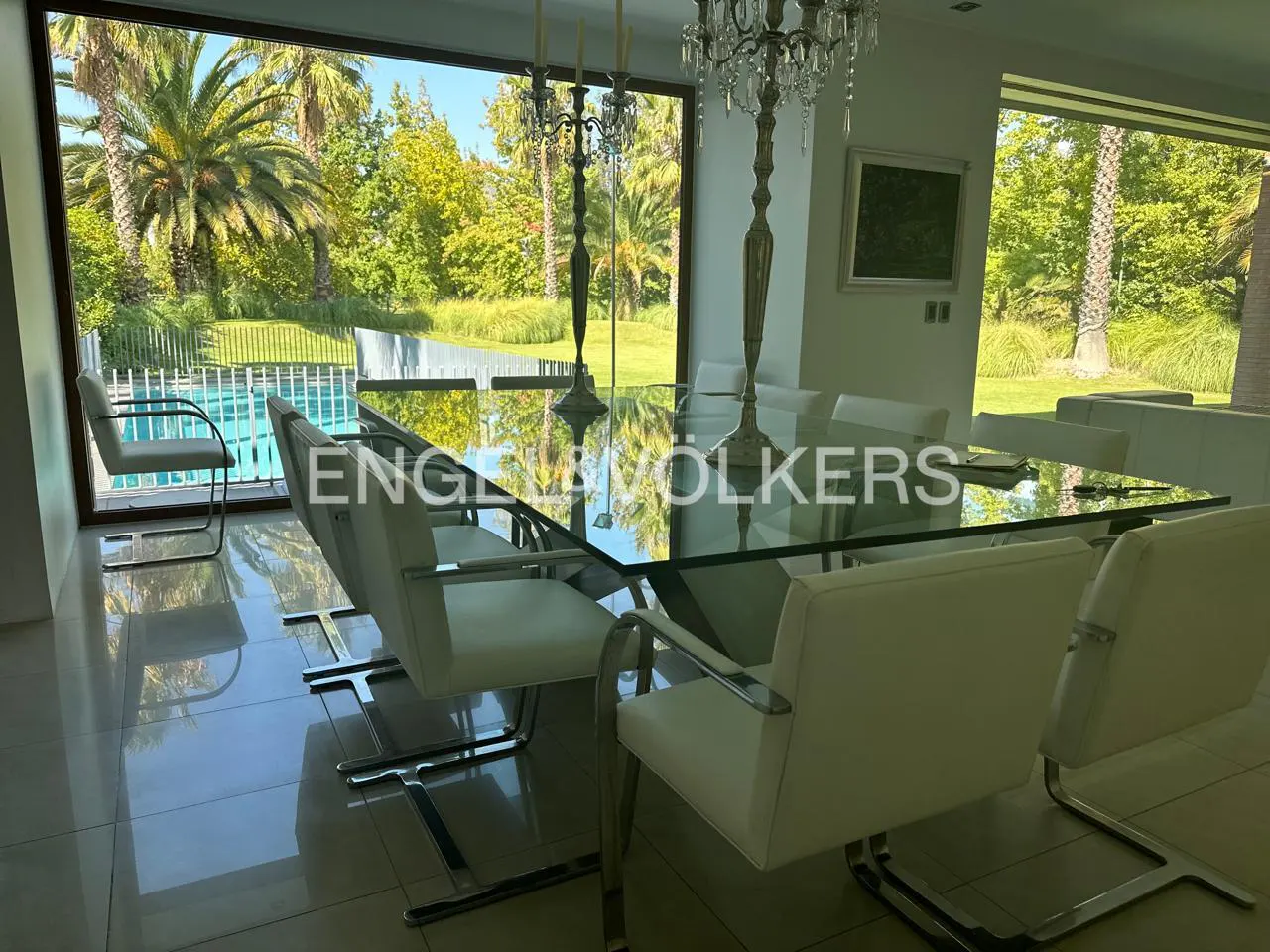 Dining room with a glass table, white chairs, and a view of a pool and palm trees through a large window. Chandeliers and candlesticks adorn the table.