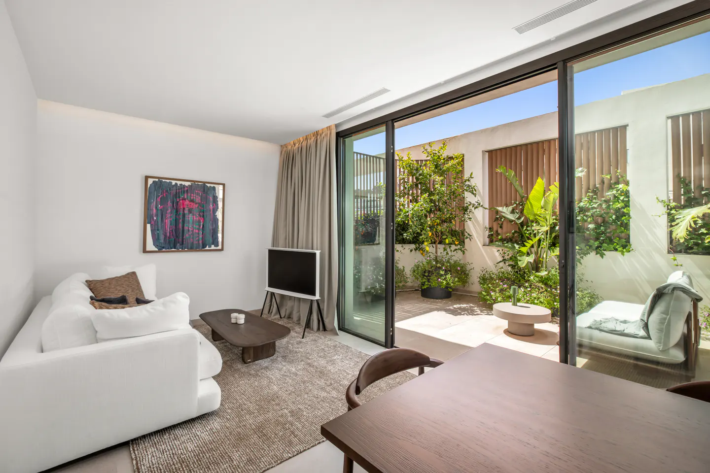 A bright living room with a white sofa, abstract art, and a TV. Sliding glass doors open to a patio with plants and a daybed.