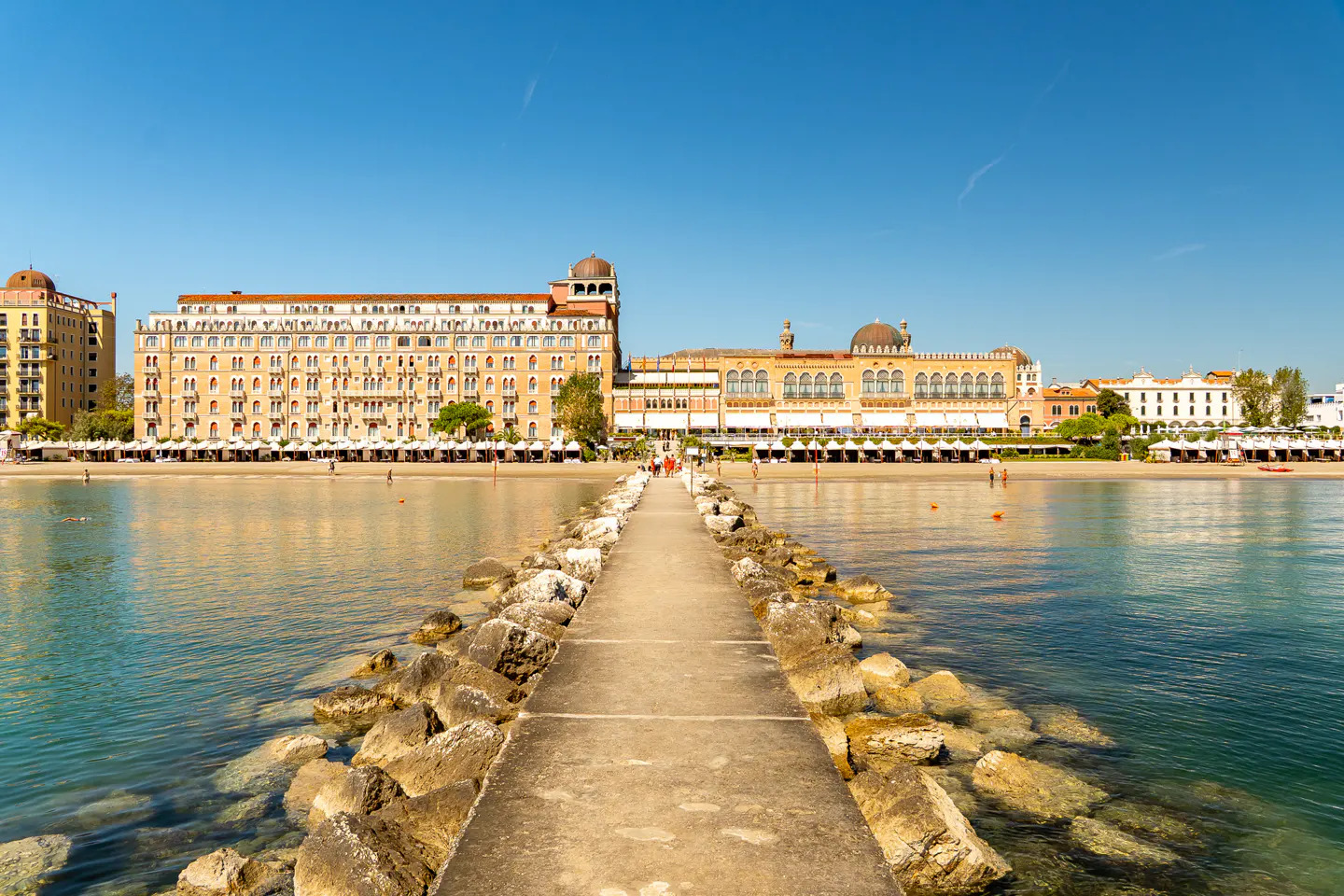 A concrete pier leads to a sandy beach with large, tan buildings under a clear blue sky.