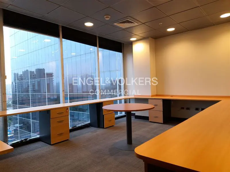 Empty office space with large windows, light wood desks, and a round table. City buildings are visible through the windows.