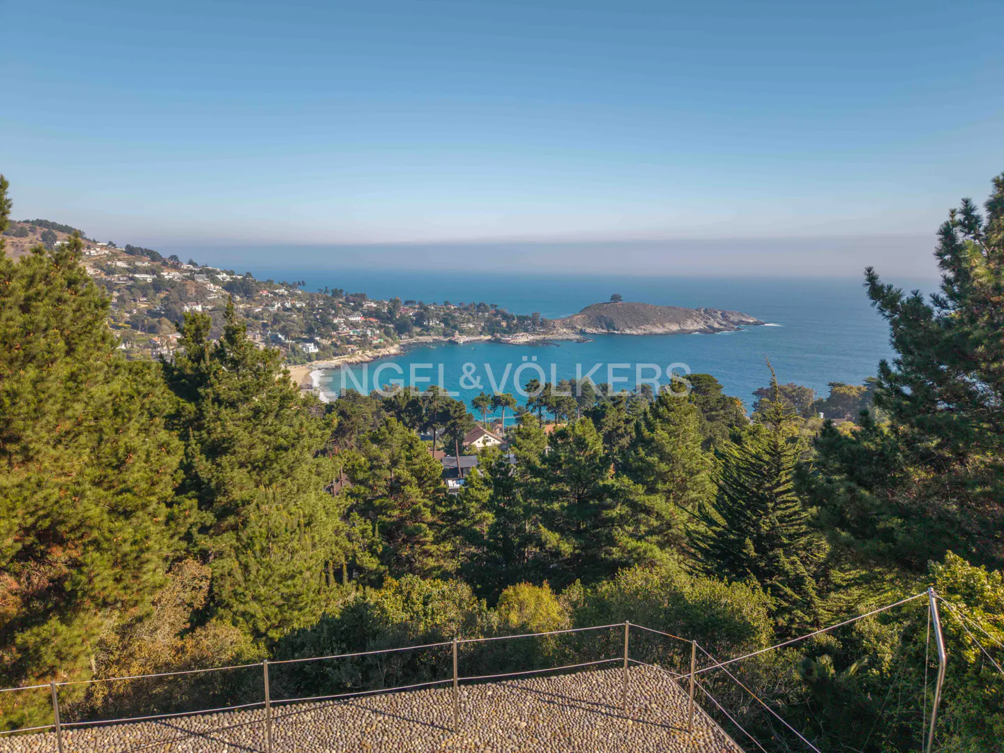 Elevated view of a lush green forest overlooking a blue coastal bay with a small town and rocky headland under a clear sky.