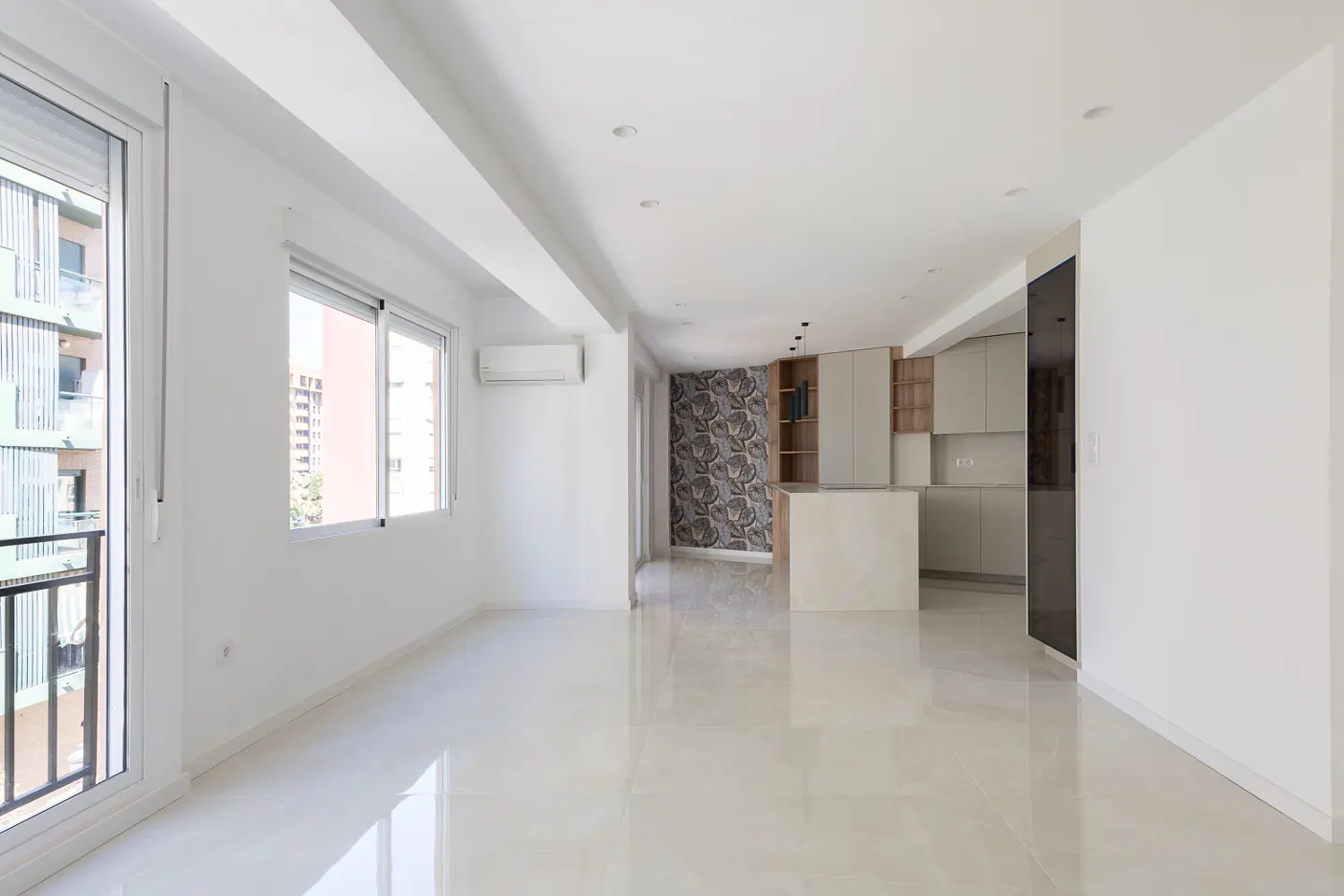 Bright, empty apartment with white walls and tile floors. A kitchen with an island is visible in the background. Balcony and windows let in natural light.
