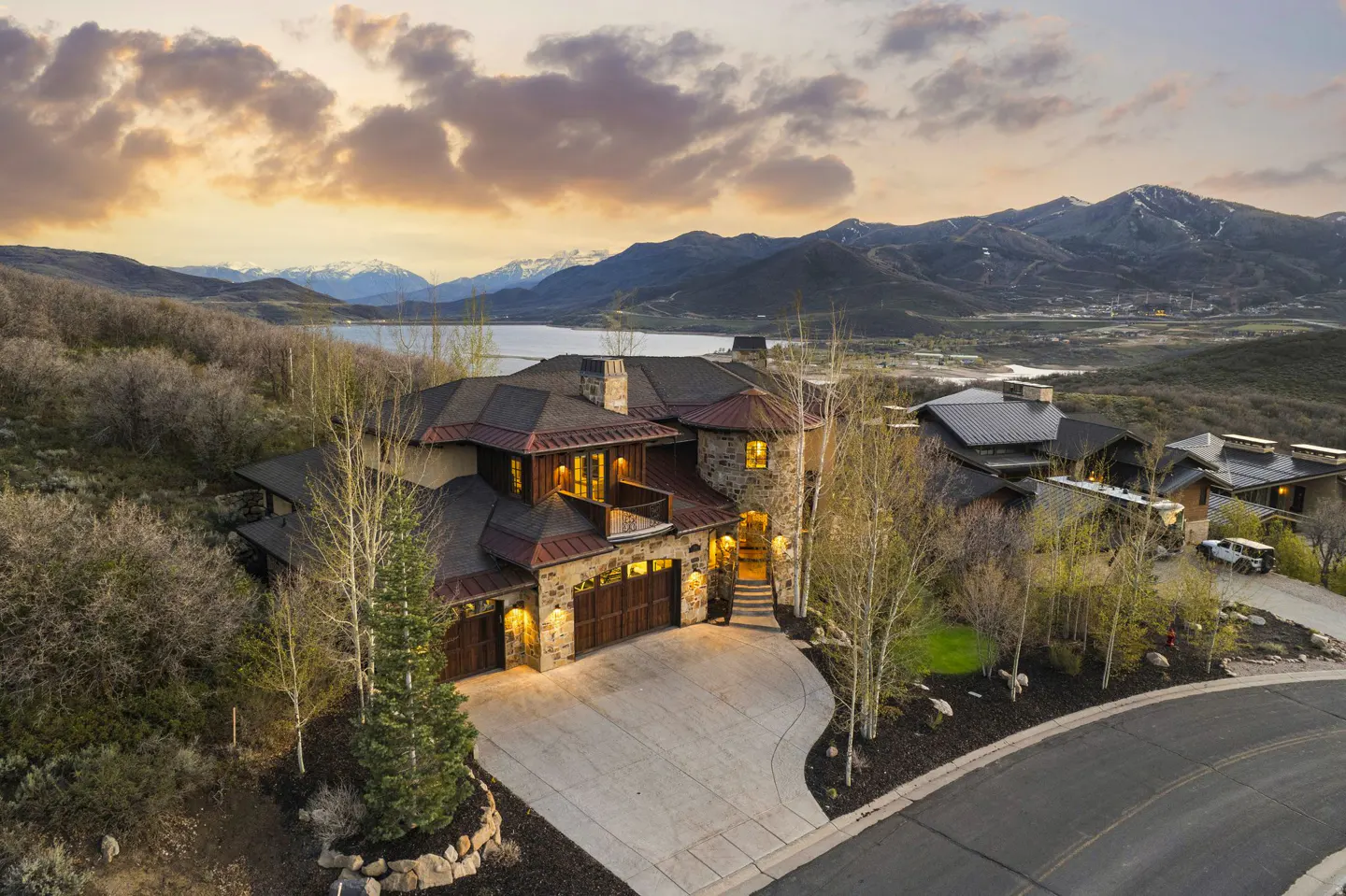 Aerial view of a stone house with a brown roof, mountains, and a lake in the background at sunset.