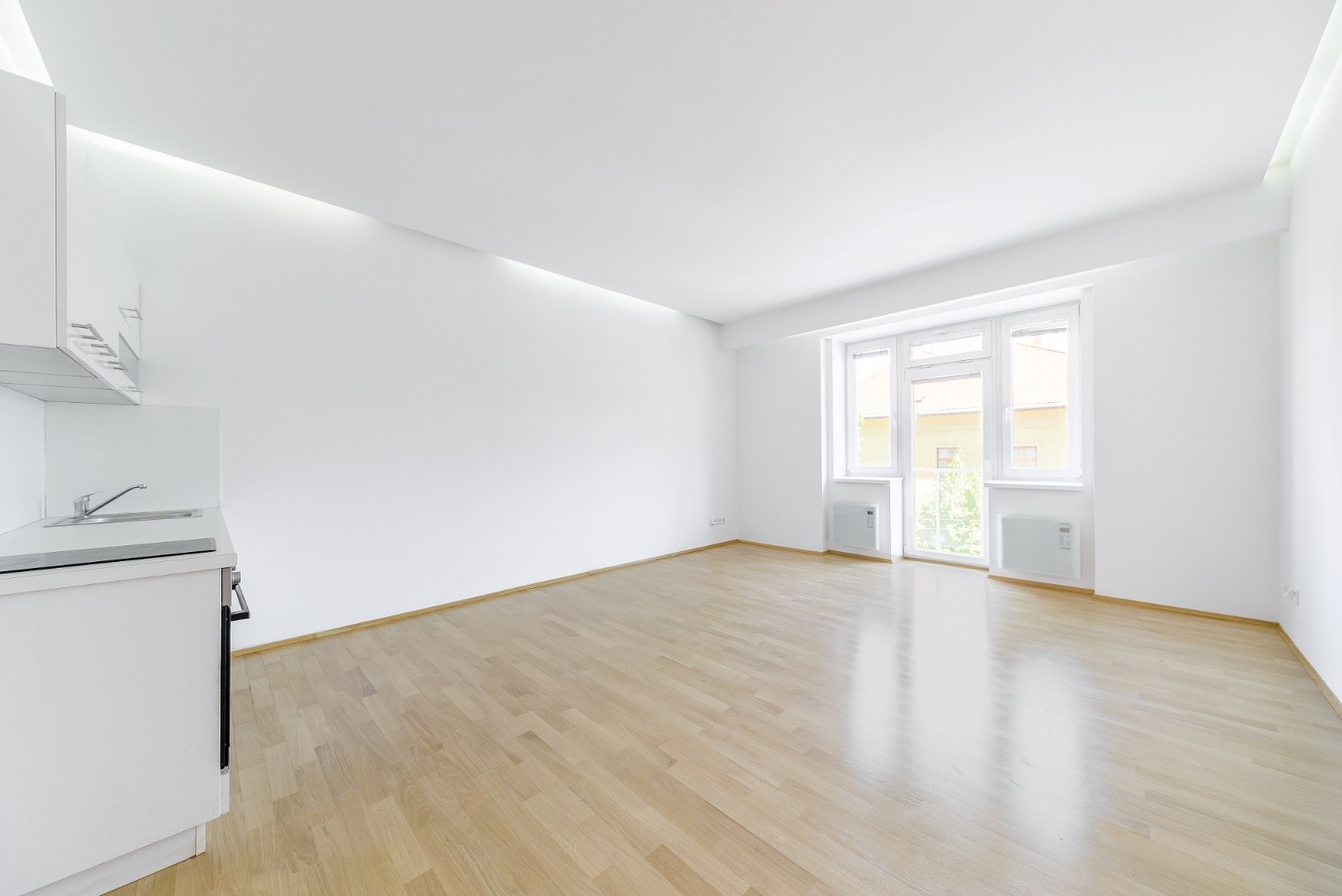 Bright, minimalist studio apartment with white walls, light wood flooring, and a small kitchen area by a glass balcony door.