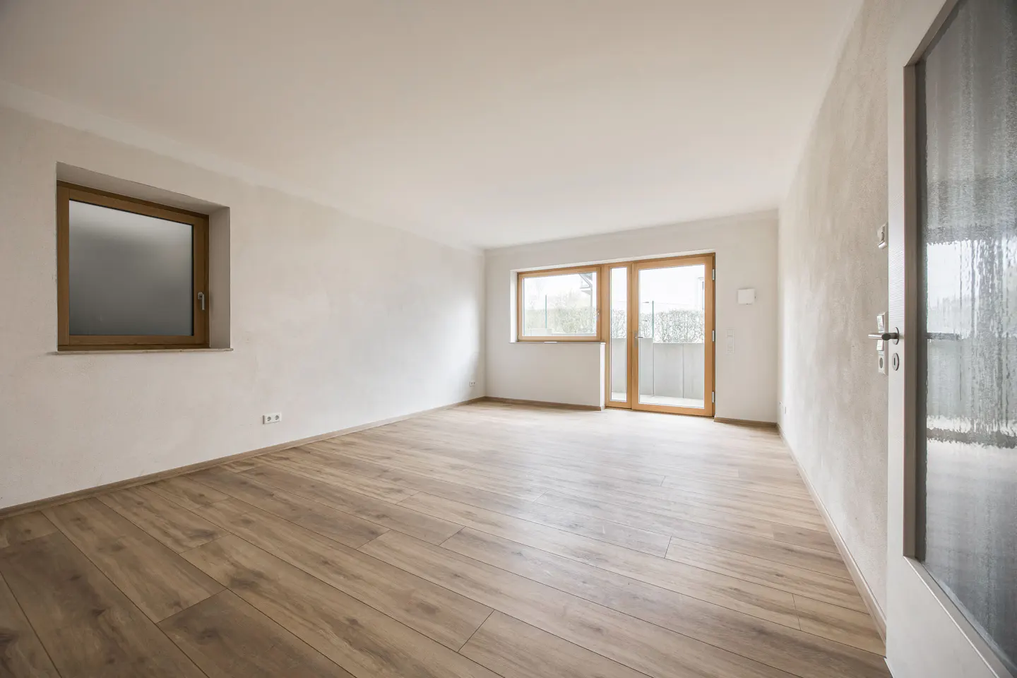 Bright, empty room with wood floors, white walls, and natural light from a window and glass doors to a balcony.