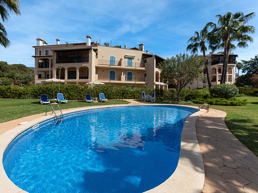 A sunny view of a blue tiled pool with lounge chairs, a tan building, and palm trees.
