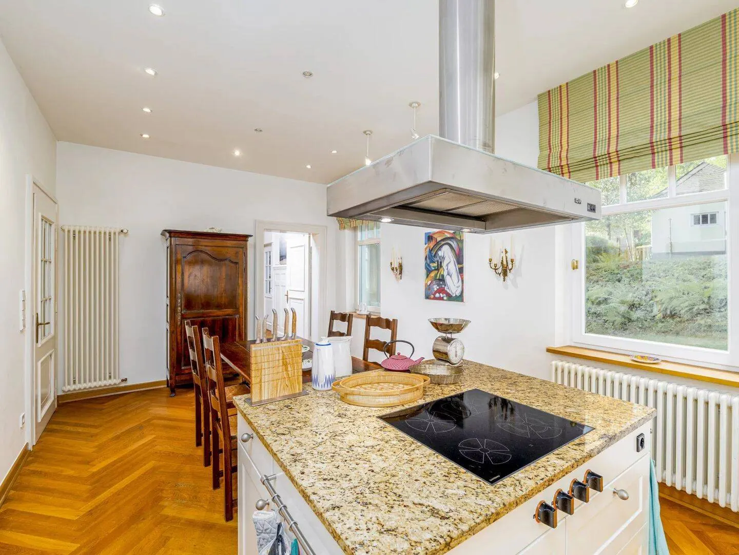 Bright kitchen with white walls, wood floors, and a granite island with a cooktop. A stainless steel range hood hangs above. A wooden table and chairs are visible.