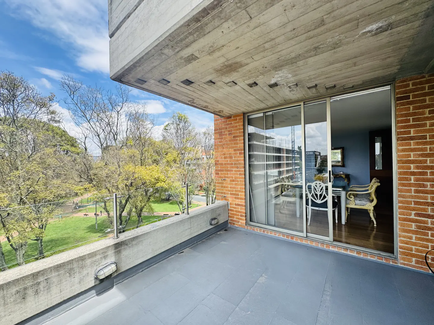 Balcony view of a brick apartment building overlooking a park with trees and a blue sky. Sliding glass doors lead to a dining area with chairs.