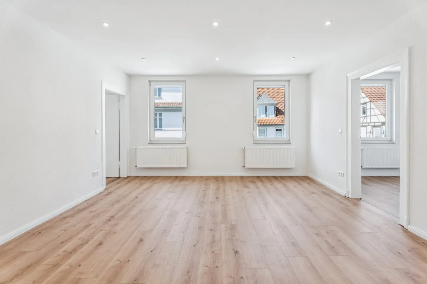 Bright, empty room with white walls, light wood floors, two windows with radiators, and doorways leading to other rooms.