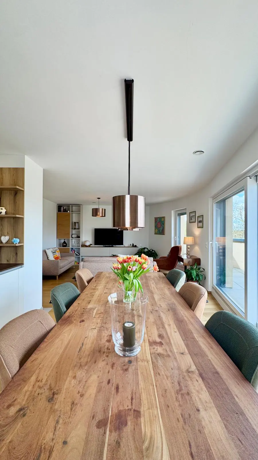 A bright, open-concept living space with a long wooden table, chairs, and a tulip centerpiece. A modern light fixture hangs above the table.