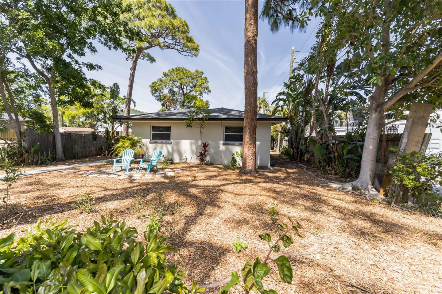 Backyard view of a one-story house with two blue chairs on a stone patio. Trees surround the yard with a mulch ground cover.