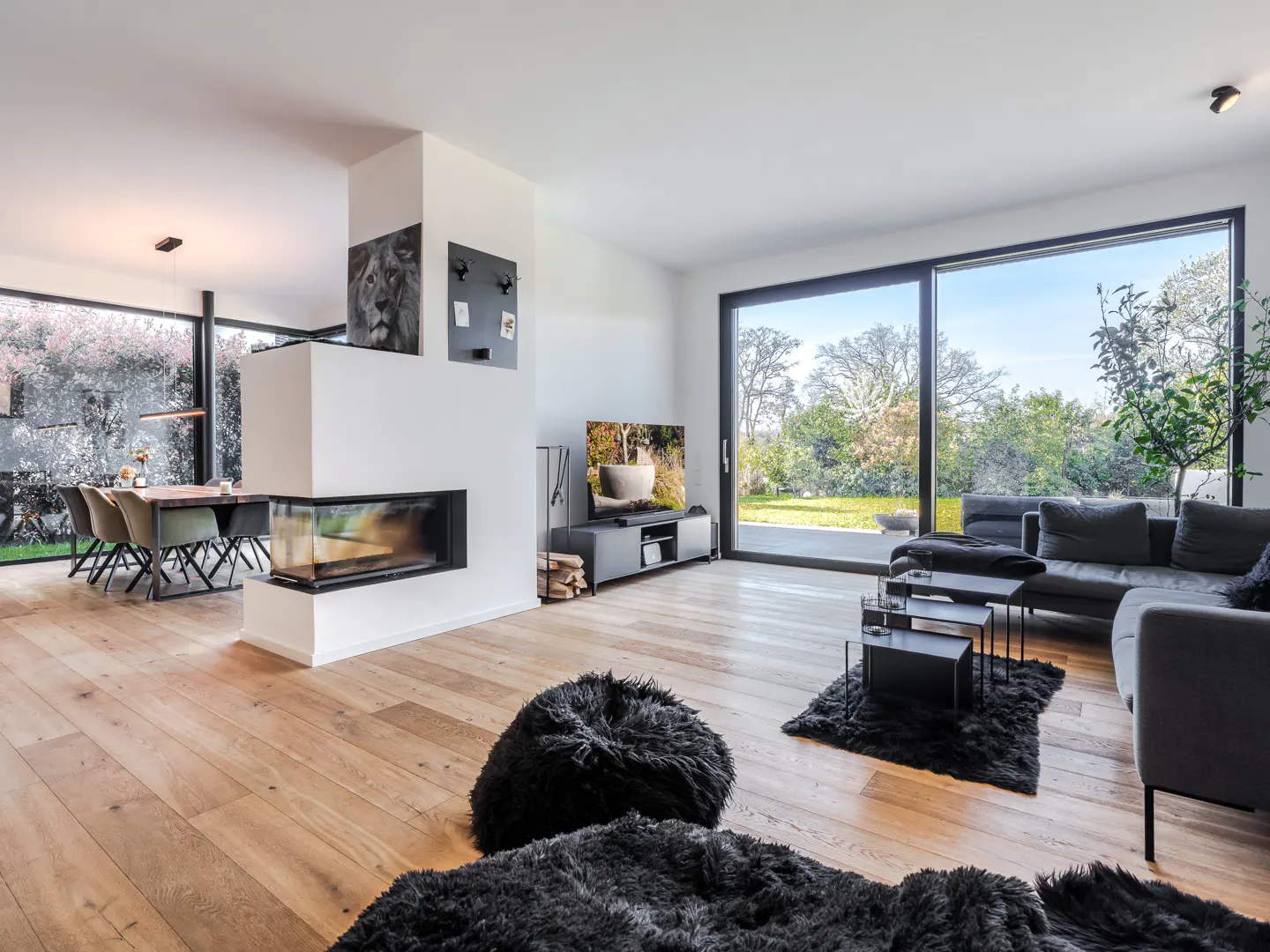 Bright living room with wood floors, a modern fireplace, and large windows overlooking a green yard. Black sofas and furry rugs add contrast.